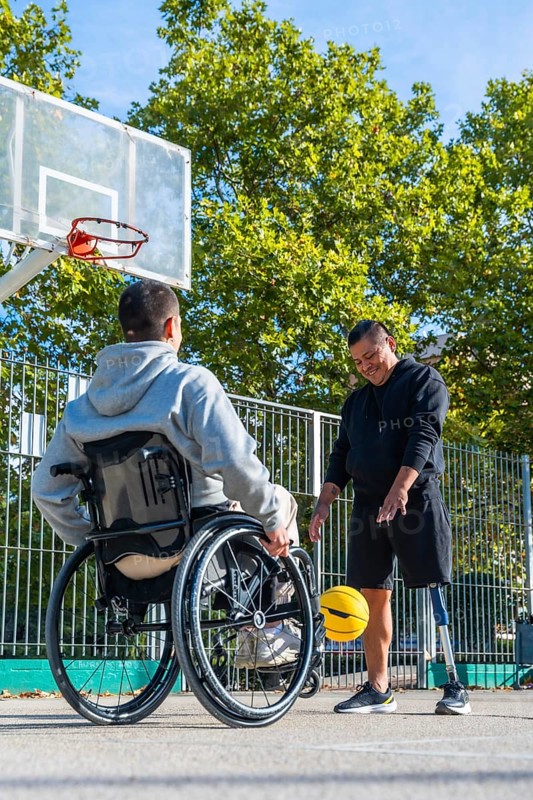 Men with a wheelchair and a prosthetic leg are happily dribbling a yellow basketball on an outdoor court, symbolizing sports inclusivity, friendship, and overcoming challenges