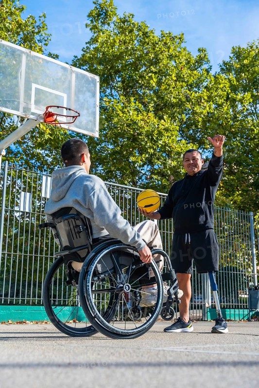 Smiling men with physical disabilities engaging in a friendly game of basketball on an outdoor court, one man using a wheelchair, the other having a prosthetic leg