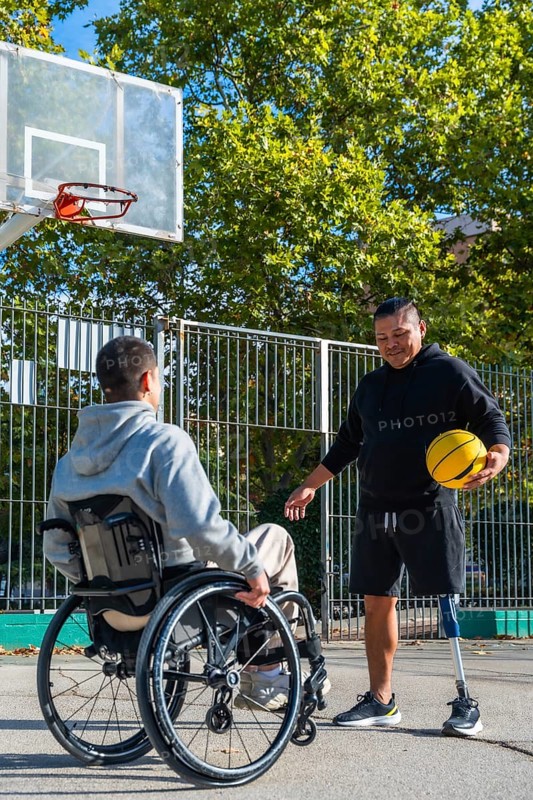 Two disabled men are enjoying a game of basketball on an outdoor court, with one man using a wheelchair and the other having a prosthetic leg, symbolizing inclusion and sport