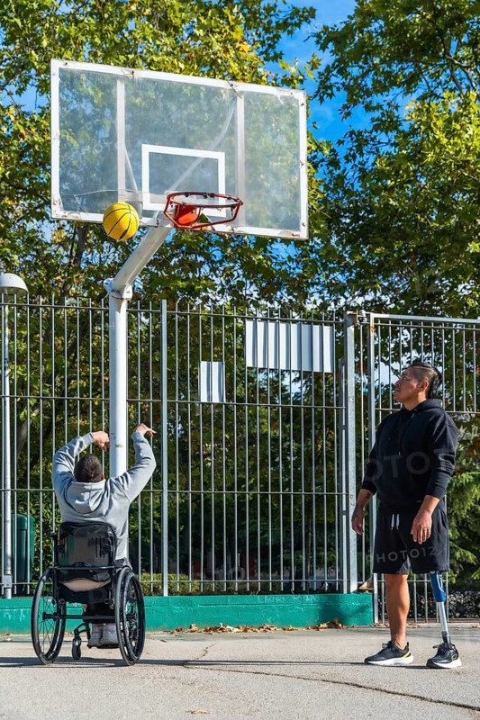 Two men play outdoor basketball, one shooting from a sports wheelchair while the other stands with a prosthetic leg, showcasing adaptive sport, teamwork and friendship on court