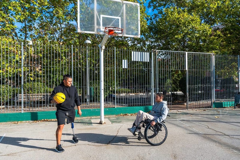 Disabled men, one using a wheelchair and the other wearing a prosthetic leg, playing basketball on an outdoor court, symbolizing inclusion and adaptive sport