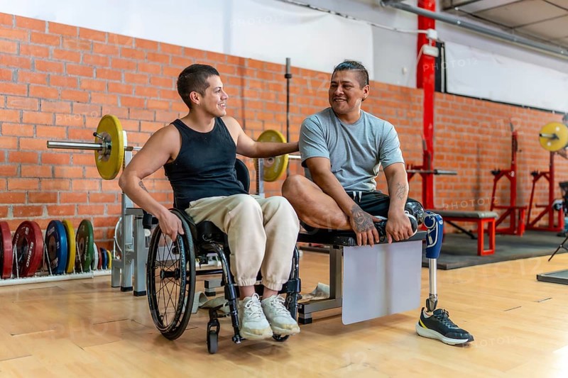 Two adaptive athletes, one in a wheelchair and one with a prosthetic leg, smiling and supporting each other while sitting in a gym, showing inclusion, determination and wellness
