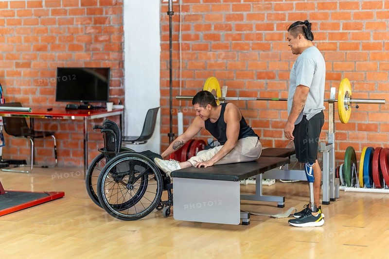 Disabled man in a wheelchair performing stretching exercises on a bench while another man with a prosthetic leg stands nearby, supporting and encouraging him during their workout session in a gym