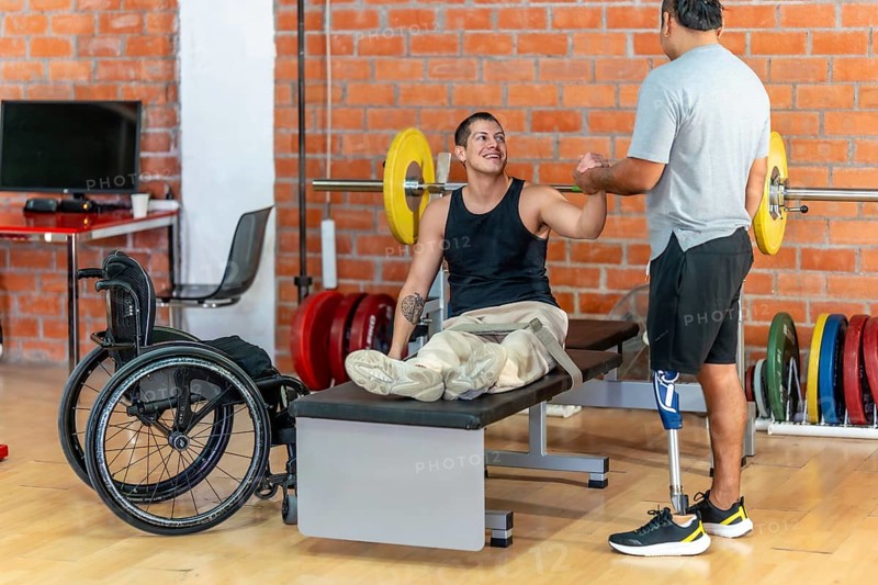 Two men with disabilities celebrate progress in a modern gym, fist bumping after a workout one using a wheelchair, the other with a prosthetic leg, smiling in solidarity and strength