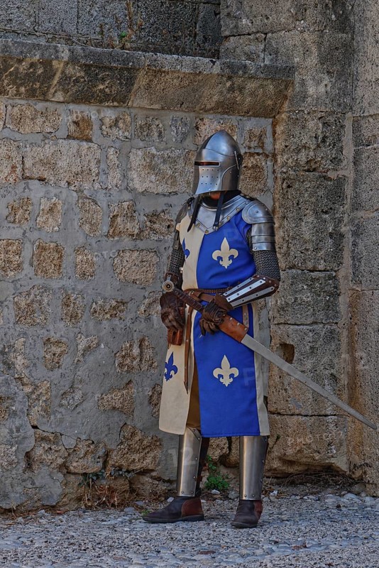 A knight in medieval armour stands with a sword in front of a stone wall, Medieval Town of Rhodes, Rhodes Island, Greece