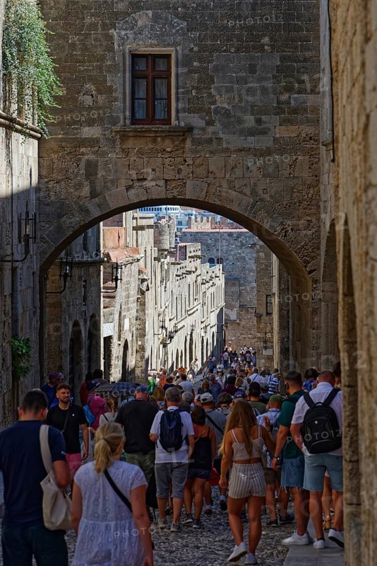 A busy historic alleyway with people walking through an alley under a stone arch, Knights Street, Medieval Town of Rhodes, Rhodes Island, Greece
