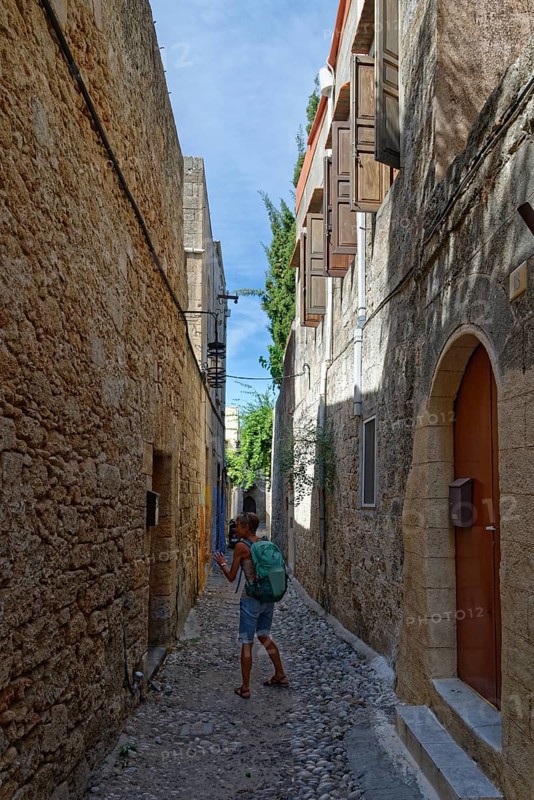 A woman with a rucksack walks through a narrow cobbled alley between historic stone houses, Medieval Town of Rhodes, Rhodes Island, Greece