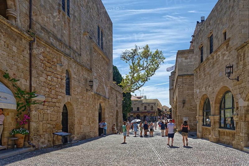 Historic old town street with cobblestones and old buildings under a blue sky full of tourists, Alexandrou Square, Medieval Town of Rhodes, Rhodes Island, Greece