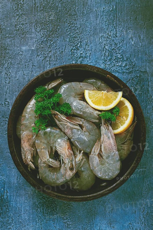 Fresh raw white-footed shrimp, tiger prawns vannamey, on a clay plate, with lemon and herbs, on a blue background