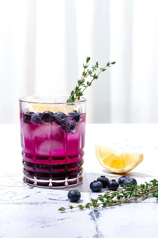 Refreshing blueberry and thyme drink served with lemon in a clear glass on a light surface