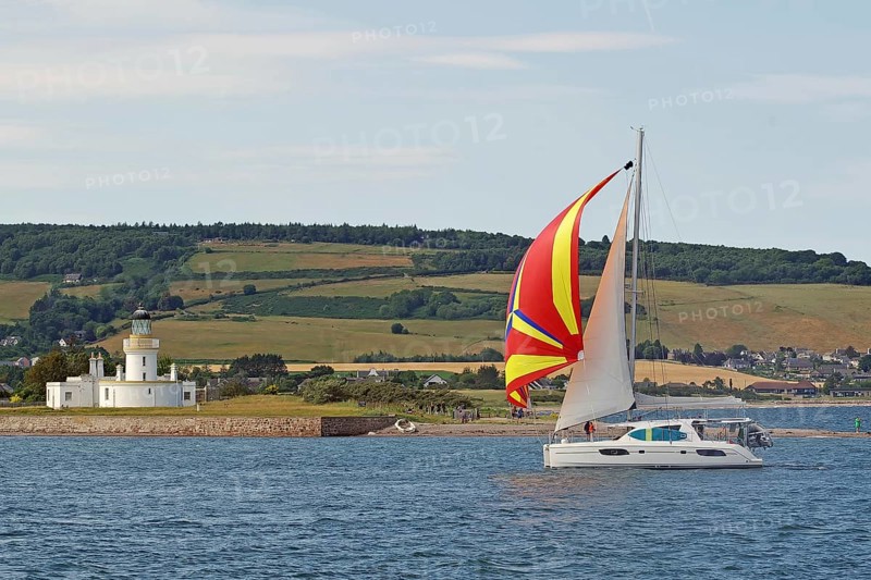 A sailing boat with a colourful sail sails near a lighthouse on the coast, Moray Firth, Inverness, Scotland, Great Britain