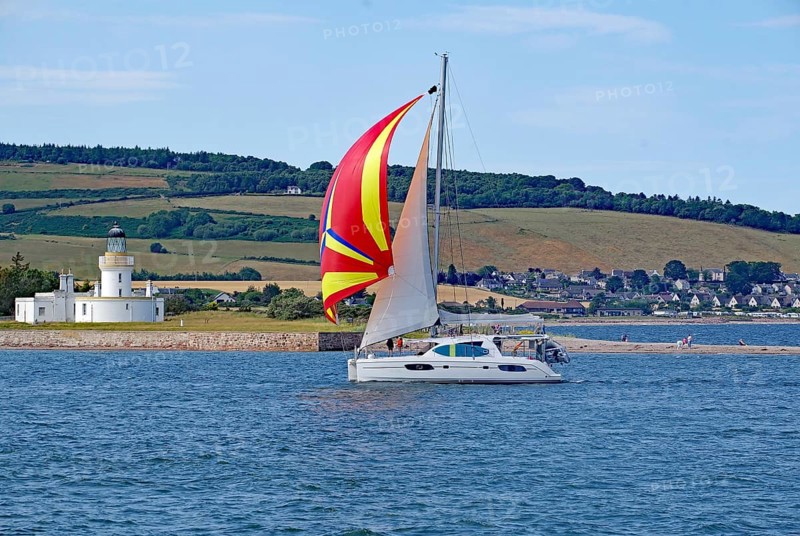 A sailing boat with a coloured sail sails near a lighthouse in front of a green landscape, Moray Firth, Inverness, Scotland, Great Britain
