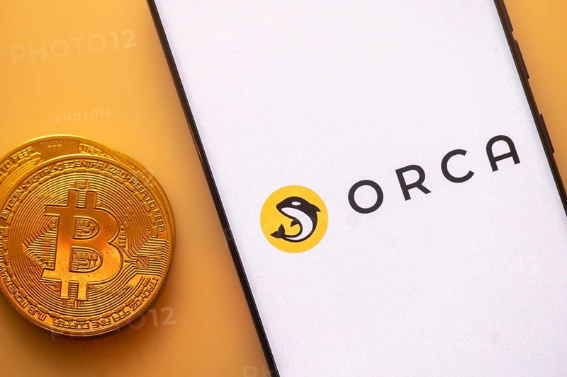 Bitcoin coin beside smartphone with Orca logo on a white screen