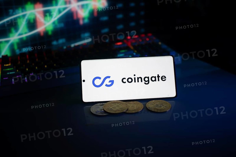 Phone with Coingate logo, set against a background of trading monitor and coins
