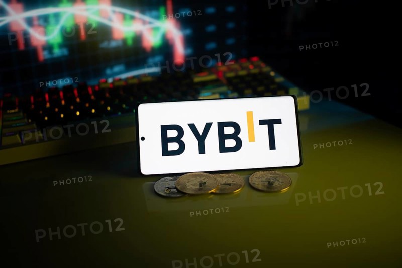 Bybit logo displayed on a phone, foregrounding trading charts and coins