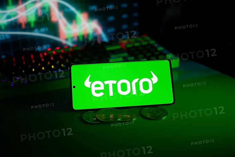 EToro logo on phone with coins and stock chart backdrop, evoking digital trading atmosphere