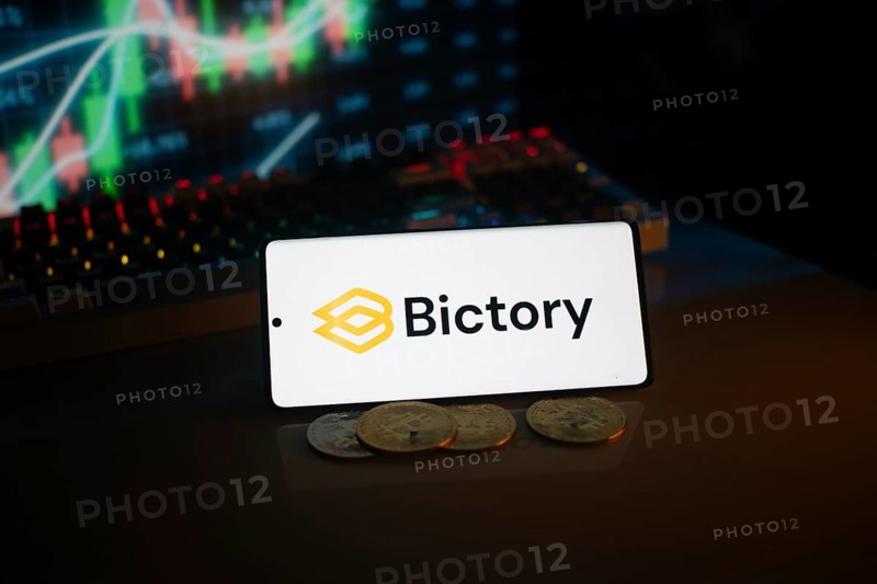 Bictory logo on smartphone amidst coins and stock chart, conveying digital and financial themes