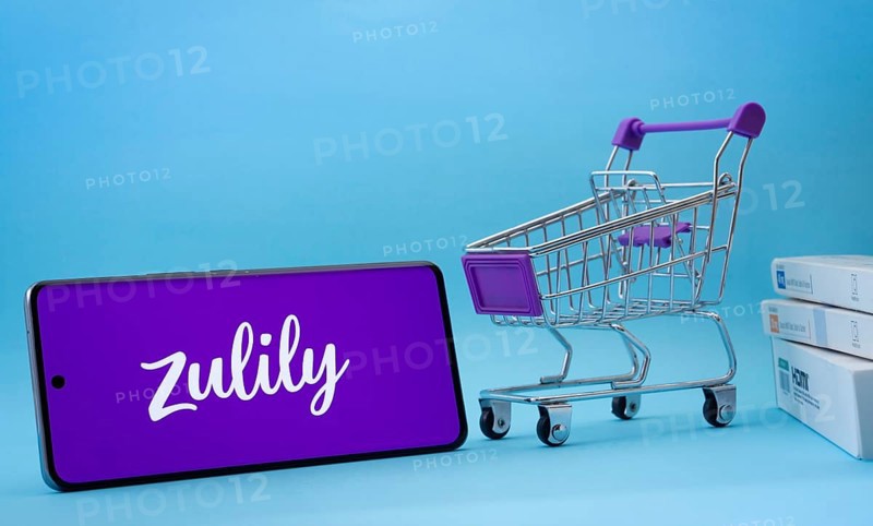 Dhaka, Bangladesh- 03 July 2025: Smartphone with Zulily logo next to a small shopping cart and boxes against a blue background