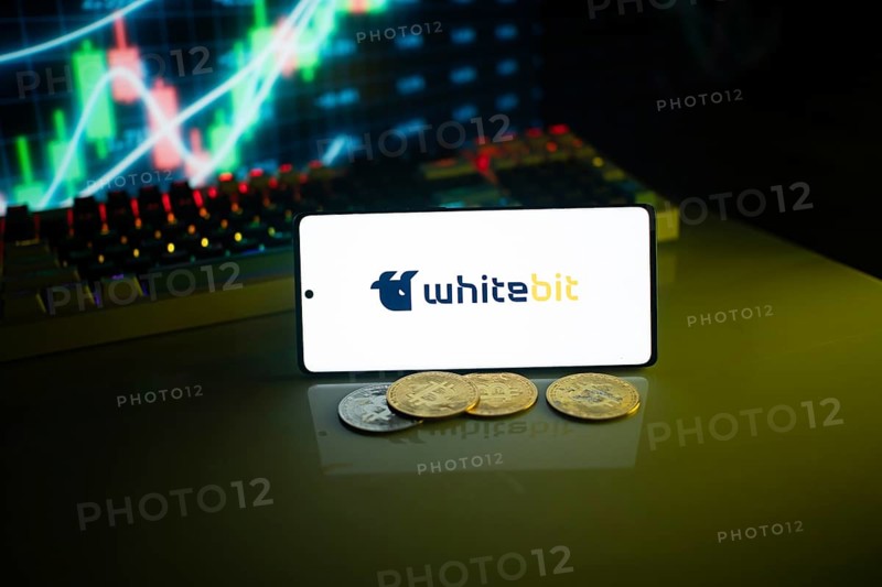 Whitebit displayed on a smartphone with coins and financial market visuals