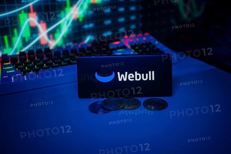 Webull depicted on a smartphone with coins and stock market indicators