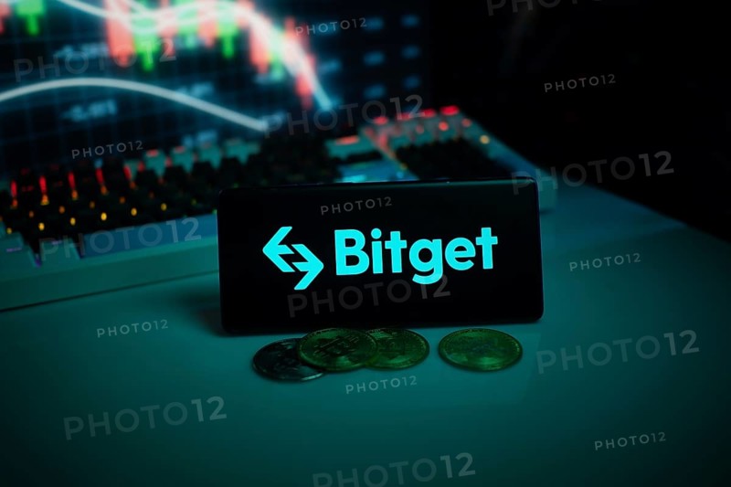 Bitget logo with coins on a desk, set against trading graphs backdrop