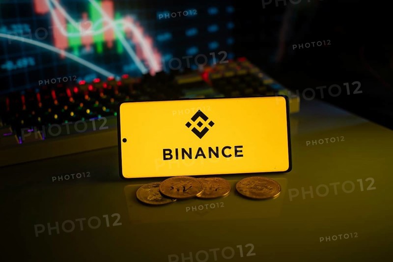 Yellow Binance logo with cryptocurrency coins on a desk, trading graphs in the backdrop