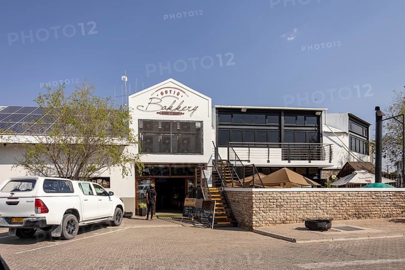 Street view of a modern building with a restaurant or café entrance in Outjo, Namibia 12 Augusto 2025: showing parked cars, outdoor seating, and clear blue sky