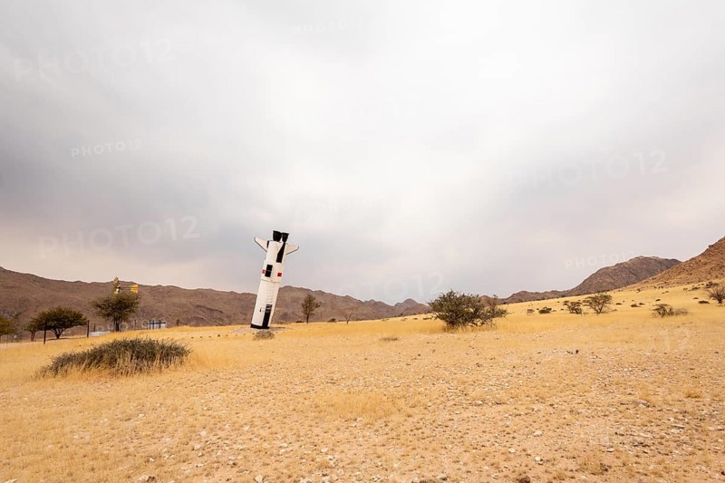 A mock Soviet rocket stands nose-first in the Solitaire, Namibian desert, surrounded by golden grasslands and rugged mountains under cloudy skies
