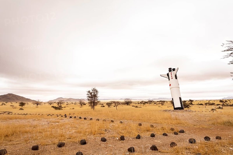A mock Soviet rocket stands nose-first in the Solitaire, Namibian desert, surrounded by golden grasslands and rugged mountains under cloudy skies
