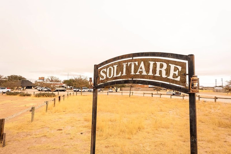 The remote desert settlement of Solitaire in Namibia features a sign displaying the town's name, set against golden sand dunes and rugged mountains