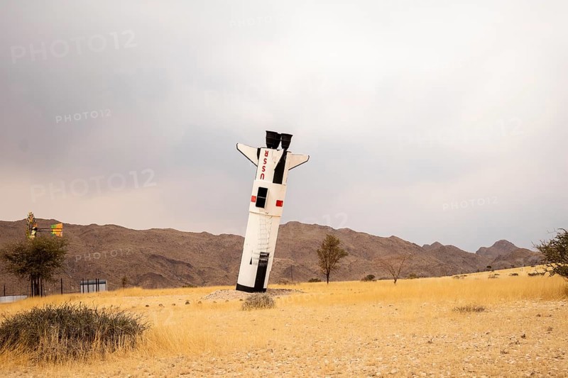A mock Soviet rocket stands nose-first in the Solitaire, Namibian desert, surrounded by golden grasslands and rugged mountains under cloudy skies
