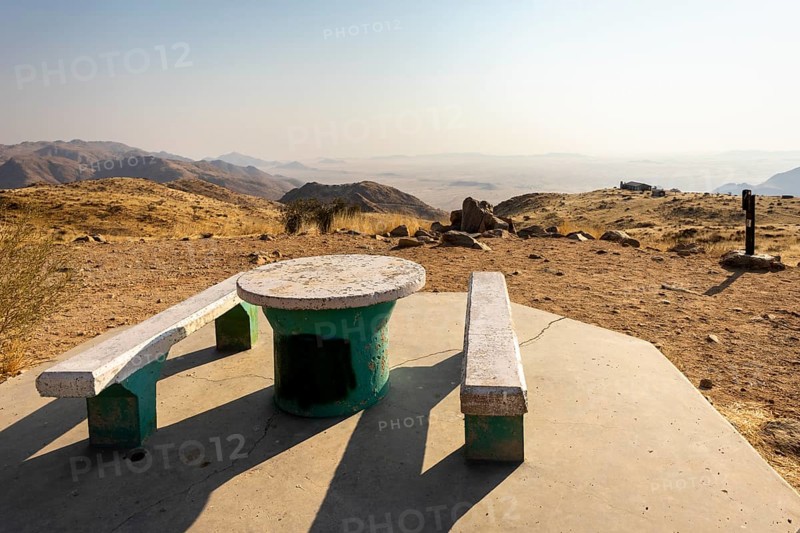 A picnic spot overlooks the vast Namib Desert from Spreetshoogte Pass, with hazy plains, rugged mountains, and a lone house on the ridge under the bright sky