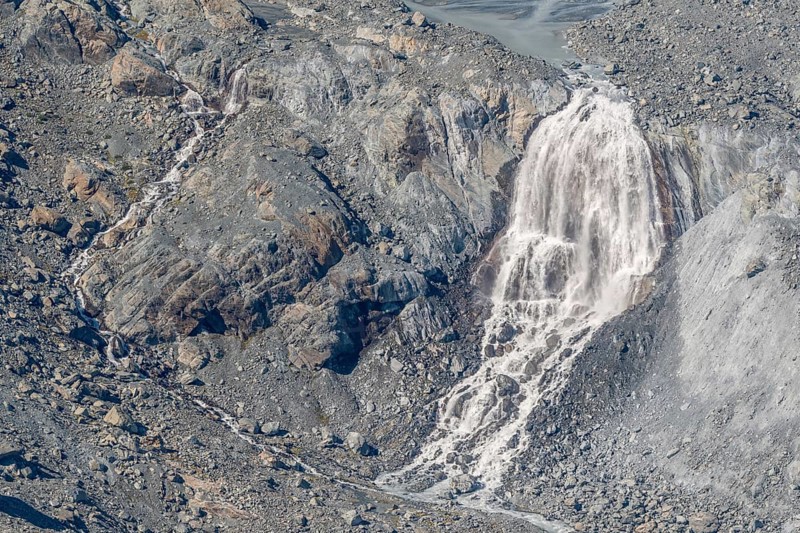 Waterfalls flow over a glacier surrounded by rocks and debris. This natural phenomenon is caused by the melting of ice, which can be observed on sunny days