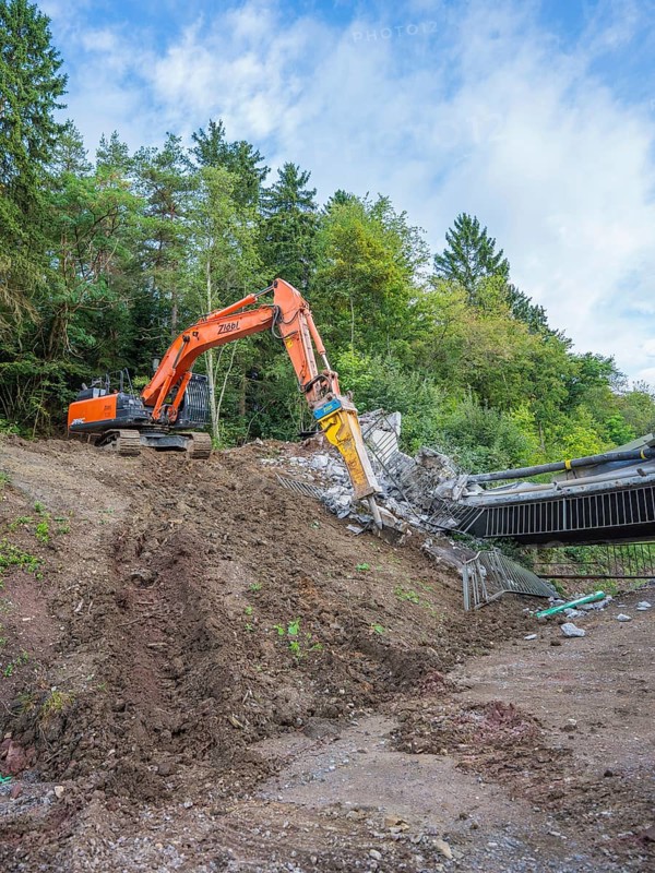 An orange excavator works in a wooded area surrounded by trees and blue sky, bridge demolition, Calw, Germany
