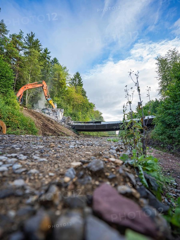 Wide angle shot of an excavator on a gravel road demolishing a bridge, surrounded by lush greenery and cloudy sky, bridge demolition, Calw, Germany