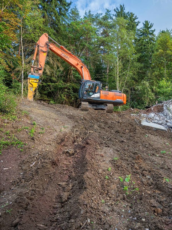 An excavator working on a pile of earth in a wooded area with a clear sky, bridge demolition, Calw, Germany