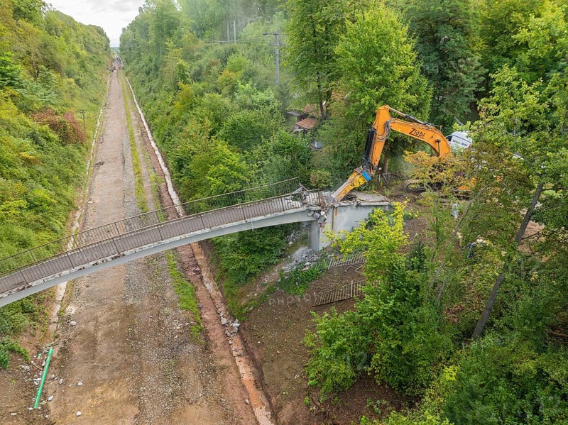 Bird's eye view of an excavator demolishing a bridge in the middle of the forest, with an empty road below, bridge demolition, Calw, Germany
