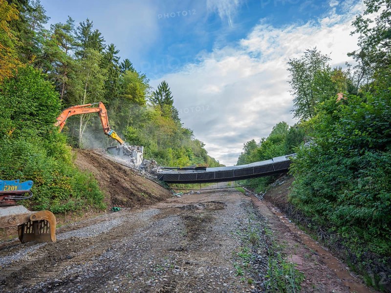 Two excavators tearing down a bridge on a gravel road, flanked by forest and a cloudy sky, bridge demolition, Calw, Germany