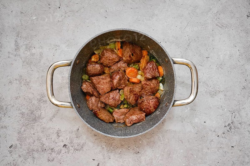 Tender pieces of beef are simmering with a mix of carrots, celery, and onions in a cooking pot. The meal is being prepared on a neutral-toned countertop in a contemporary kitchen setting