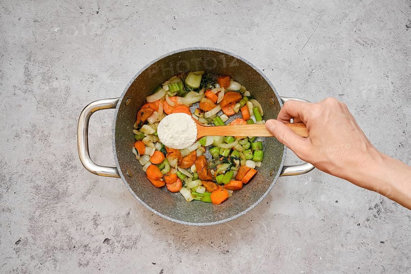 Freshly chopped carrots, celery, and onions are fried in a pot. A hand is sprinkling flour over the colourful mixture, indicating the preparation of a savory dish in a home kitchen