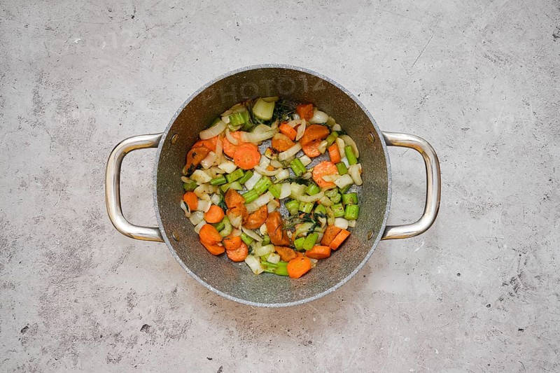 Chopped carrots, celery, and onion are frying in a pot, creating a vibrant vegetable base mixture on kitchen tabletop