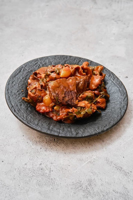 A hearty meat stew is presented on an elegant black plate. The dish features tender chunks of beef cooked with assorted colorful vegetables in a savory mushroom sauce, highlighting rich aromas and flavors