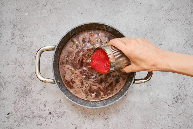 A hand pours diced tomatoes from a can into a pot filled with a rich, simmering beef stew