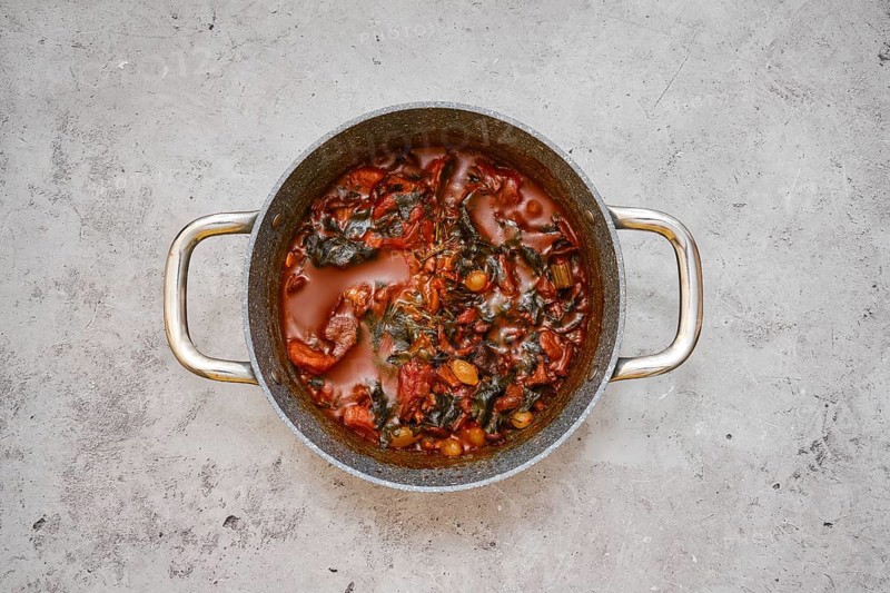 A large pot filled with a flavourful beef and vegetable stew on a gray kitchen countertop. Ingredients like tomatoes and leafy greens are visible, suggesting a healthy home-cooked meal
