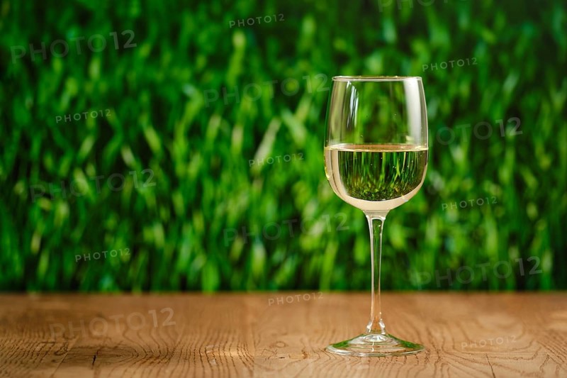 A clear glass filled with white wine on a wooden table. The backdrop features lush green grass, creating a refreshing atmosphere perfect for relaxation