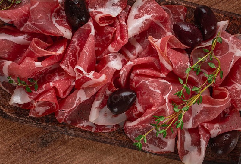 Freshly cut dried meat, served with olives and herbs, on a wooden chopping board, no people