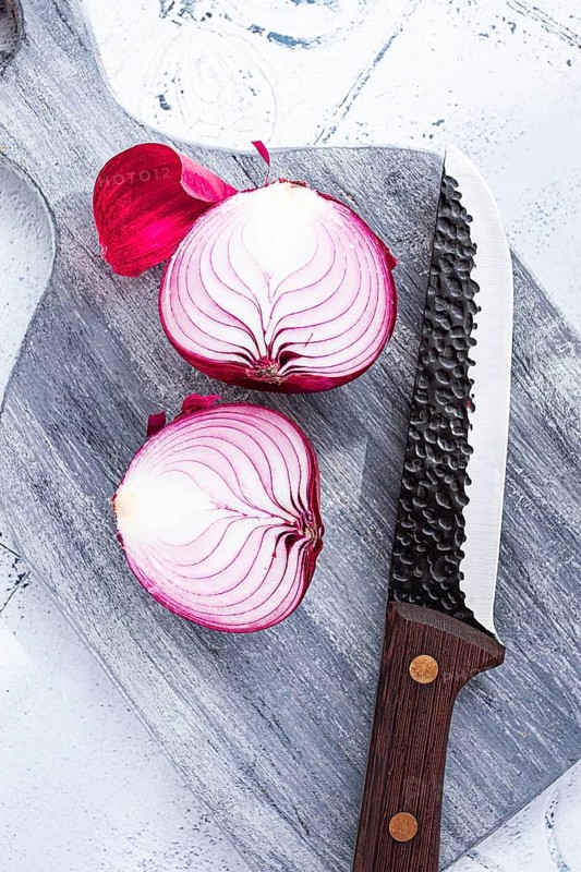 A vibrant red onion is cut in half, on a rustic wooden chopping board beside a sharp knife, ready for cooking