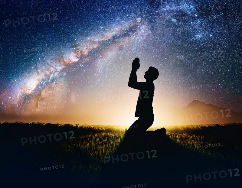 A man kneels in peaceful reflection under the vibrant Milky Way galaxy at sunset in an open natural setting, making prays, AI generated