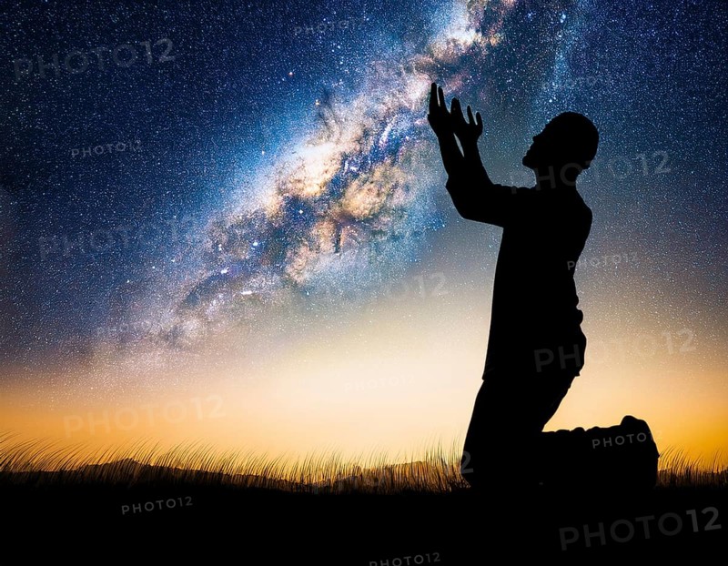 A man kneels in peaceful reflection under the vibrant Milky Way galaxy at sunset in an open natural setting, making prays, AI generated