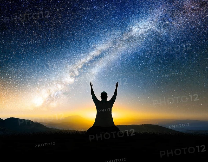 A man kneels in peaceful reflection under the vibrant Milky Way galaxy at sunset in an open natural setting, conveying awe, tranquility, and the beauty of the cosmos, AI generated